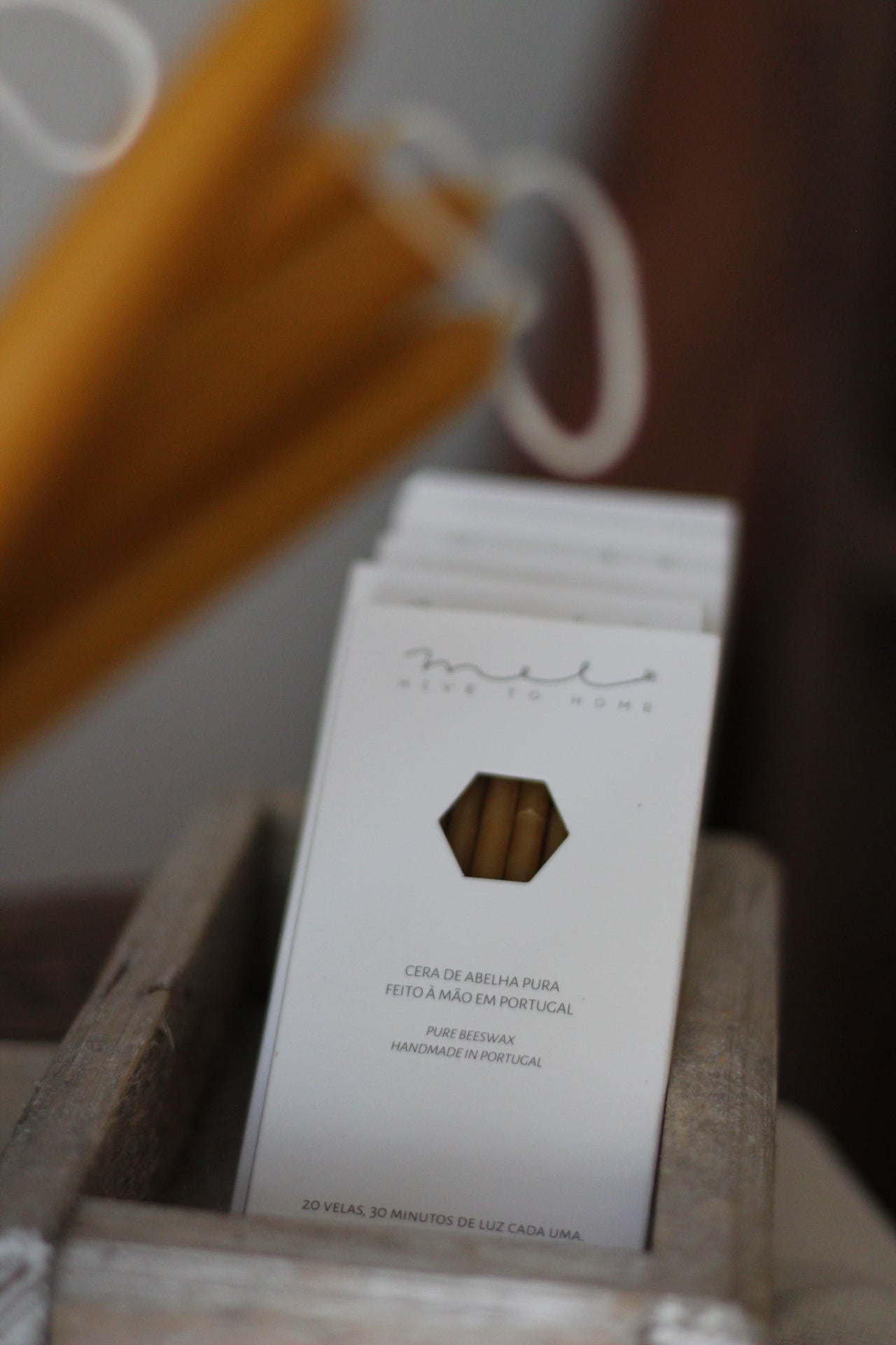 A close-up of a white box containing beeswax candles, with a small hexagonal window showing the golden-yellow beeswax candles inside. The box is resting on a wooden shelf, with a blurred background showing more candles and a white cord.
