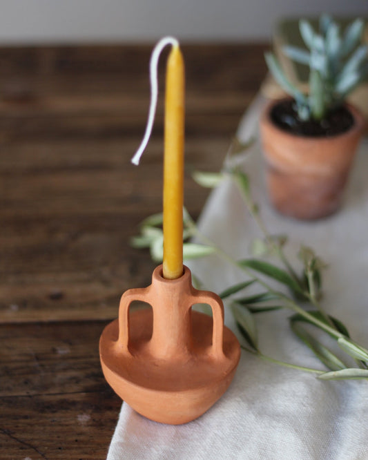 A terracotta clay candle holder with a yellow taper candle inserted, styled on a white fabric runner over a wooden table with a small potted succulent in the background.
