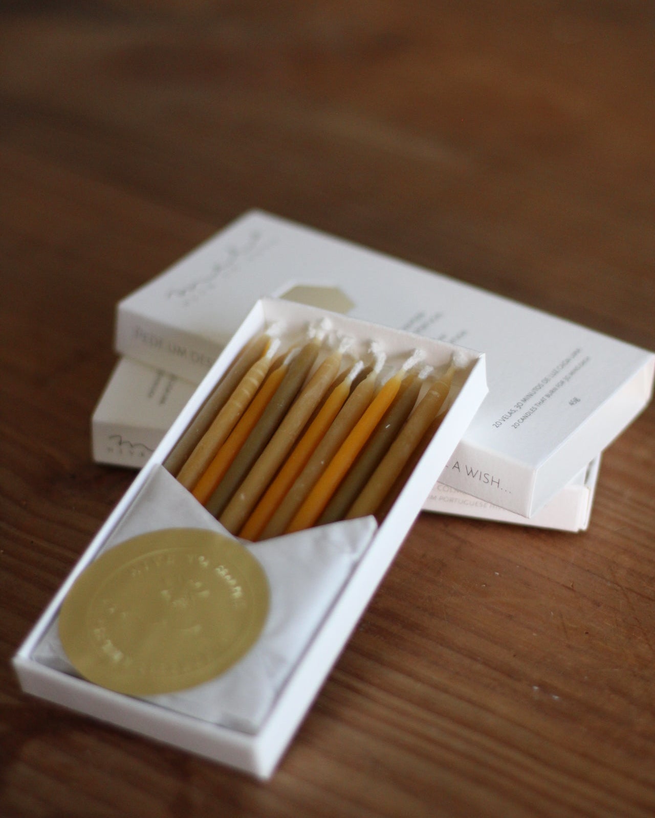 An open white box containing several yellow-orange beeswax candles, placed on a wooden surface. A gold circular seal is visible on the box lid.
