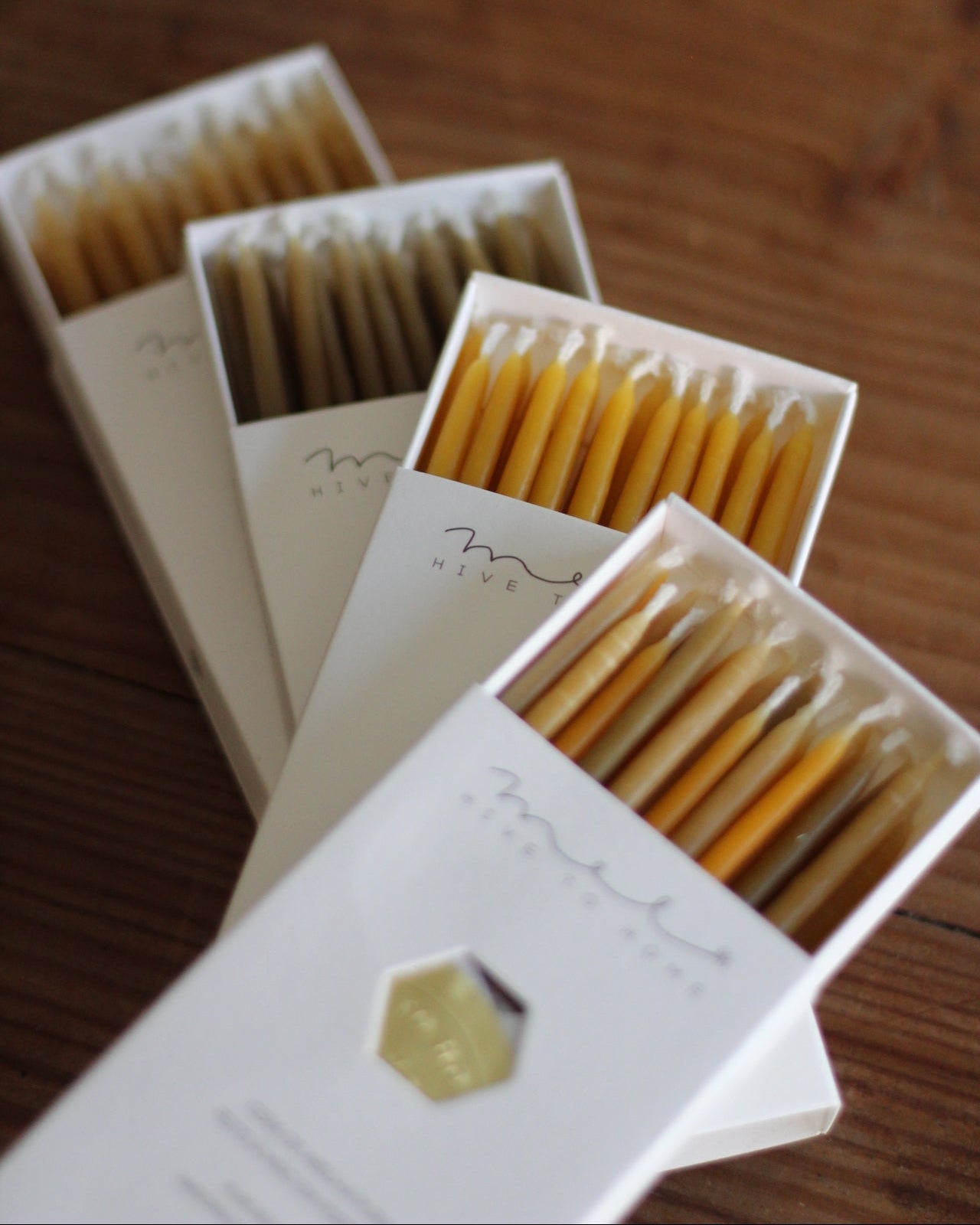 Three open white matchboxes containing beeswax candles, arranged diagonally on a wooden surface.