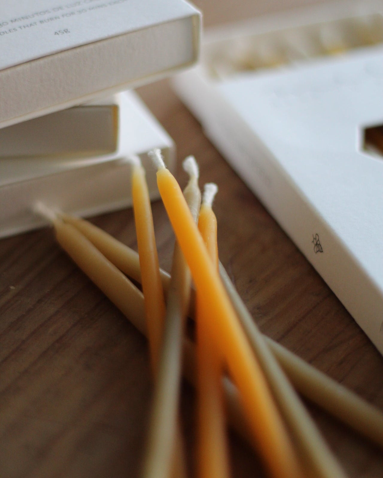 A close-up angled view of several natural beeswax candles in a warm golden-yellow color resting on a wooden surface next to white packaging boxes.