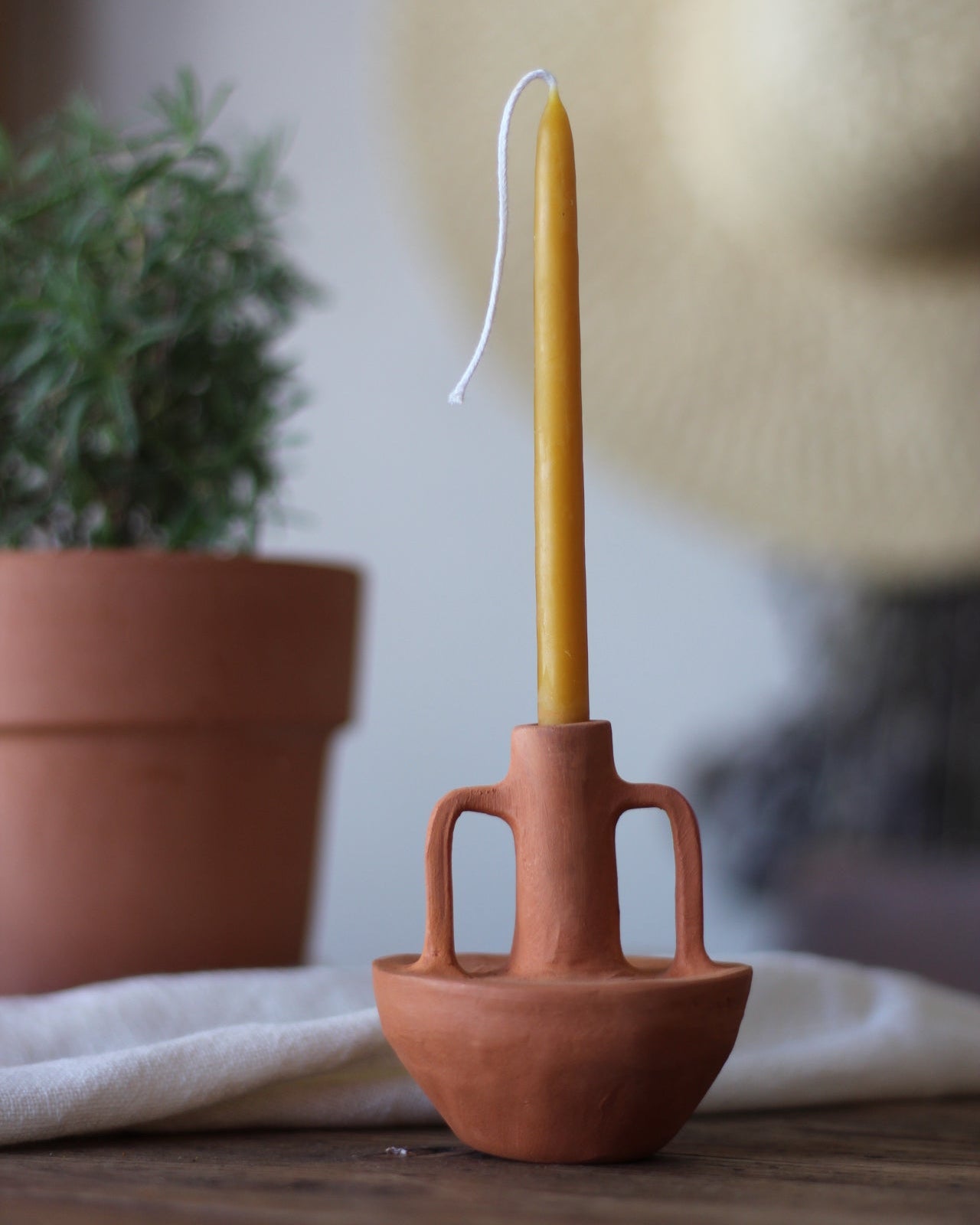 A terracotta clay candle holder with two handles holding a yellow taper beeswax candle, styled on a wooden surface with a potted plant and a woven hat in the background.