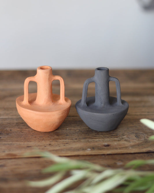 Two small ceramic vase-like vessels with looped handles sit on a rustic wooden surface against a soft light gray background. One is terracotta orange and the other is dark charcoal gray, with blurred green leaves in the foreground.