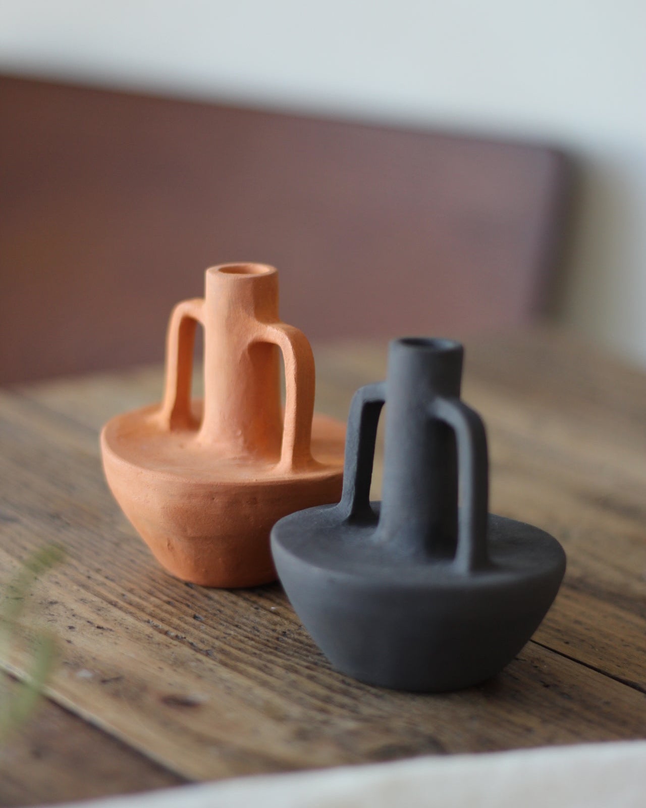 Two small ceramic vase-like candle holders with loop handles sit on a rustic wooden table. One is terracotta orange and the other is dark charcoal gray, photographed in soft natural light with a blurred indoor background.