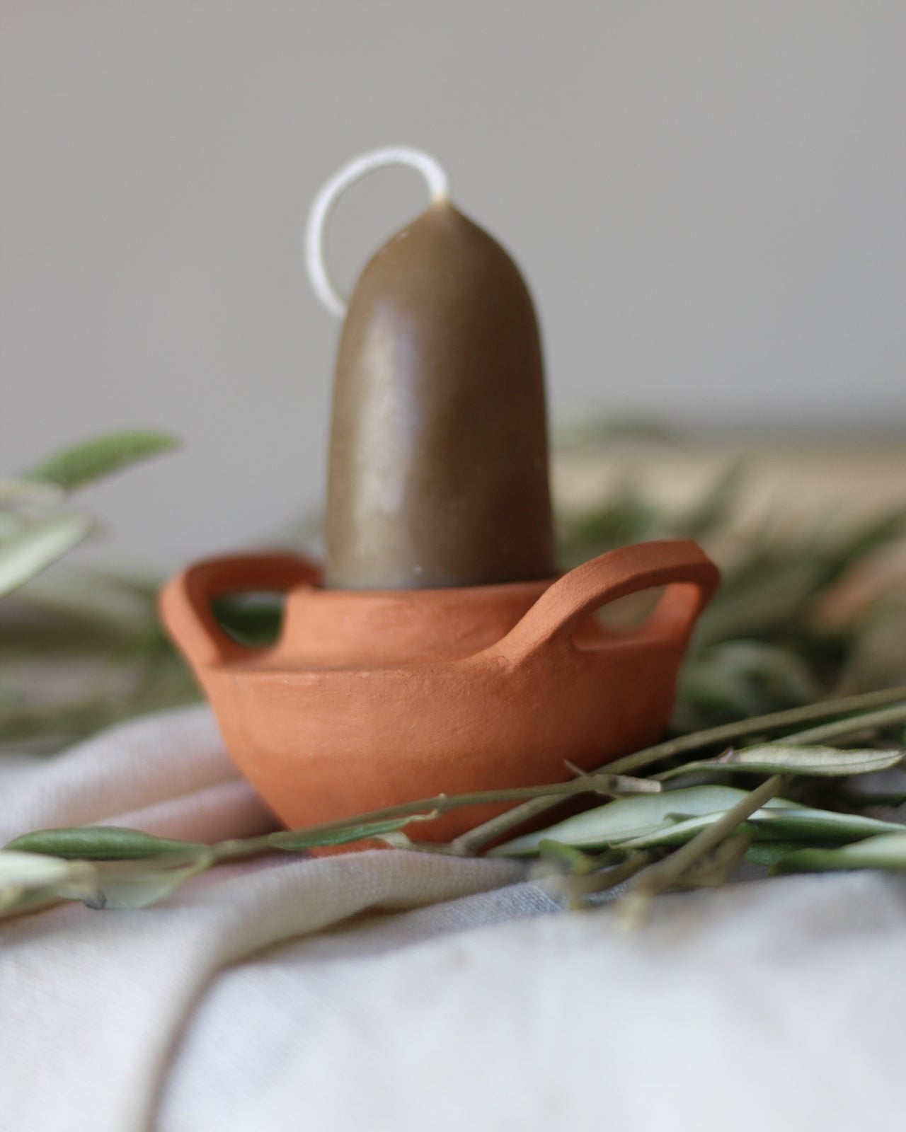 A matcha green taper candle displayed inside a terracotta-colored ceramic pot with side handles, surrounded by dried greenery on a soft white fabric surface.